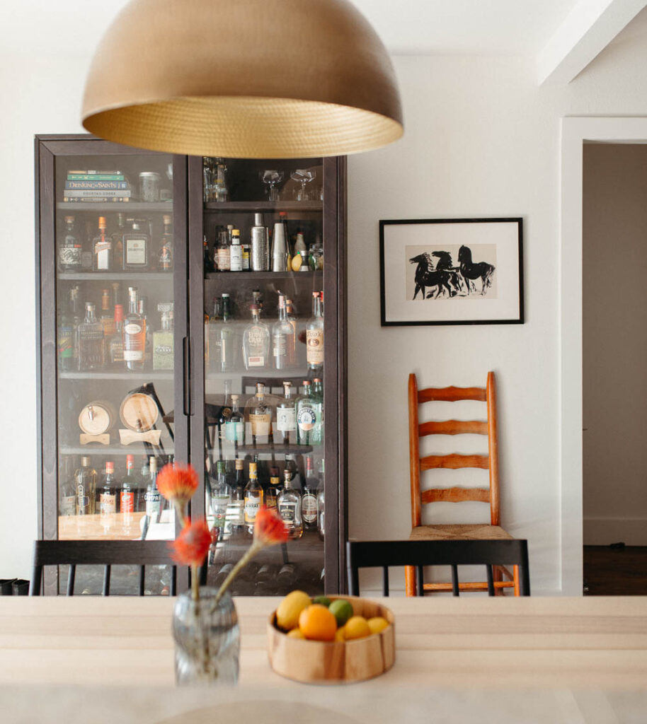 Off centered art above an antique chair next to a bar cabinet.